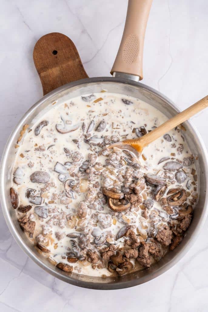 Sour cream and sauces added to a saucepan with ground beef, mushrooms, garlic and onions