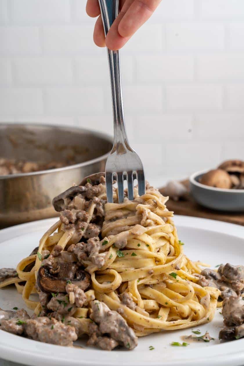 Ground beef stroganoff served on a white plate with buttered noodles and a fork inserted to swirl the noodles for serving.