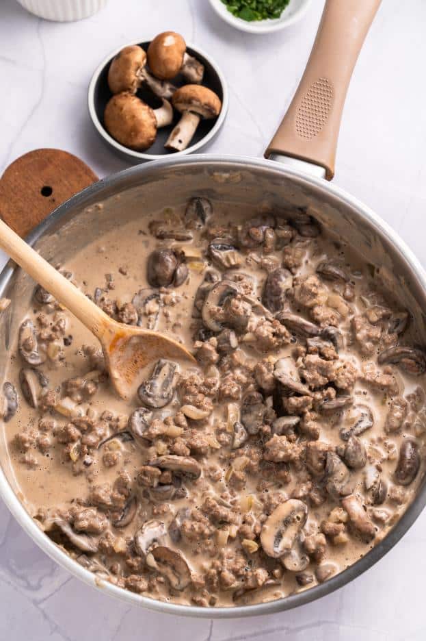 A saucepan filled with a creamy mushroom and beef sauce being stirred by a wooden spoon.
