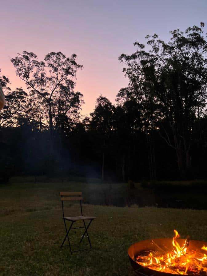Night time in the bush with a pink and purple skyline, an outdoor fire and a single chair in a paddock