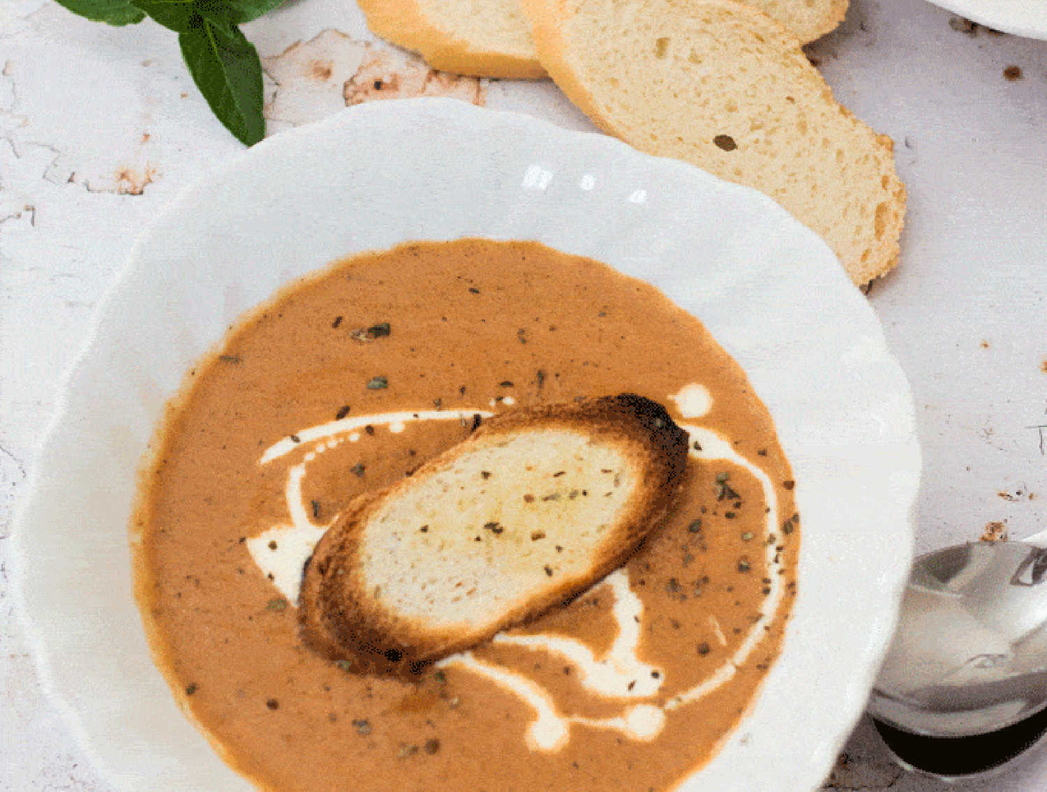 A white bowl containing tomato soup topped with a drizzle of cream and a slice of toasted baguette